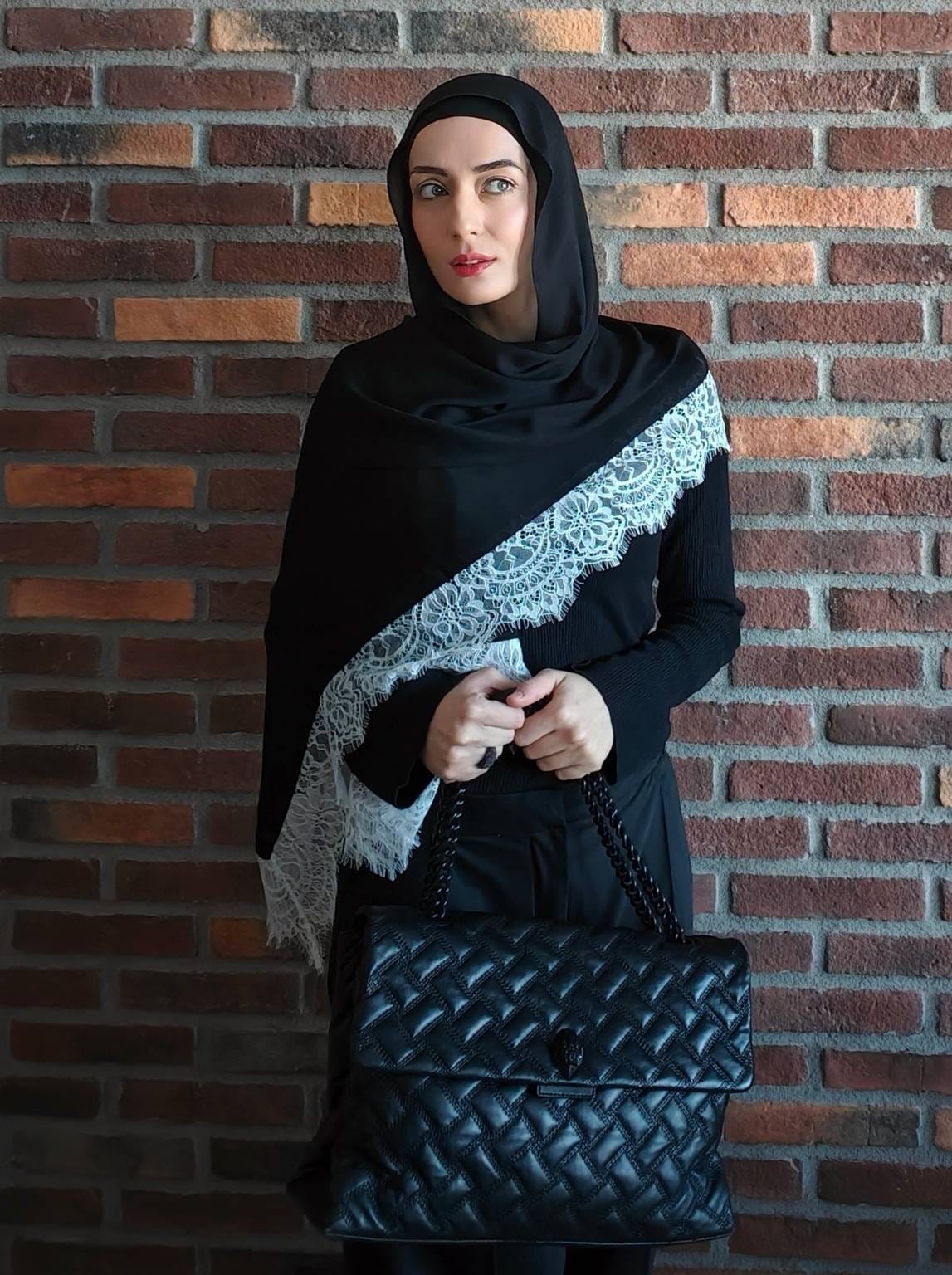 Black lace hijab draped diagonally over an all-black modest outfit against a brick wall