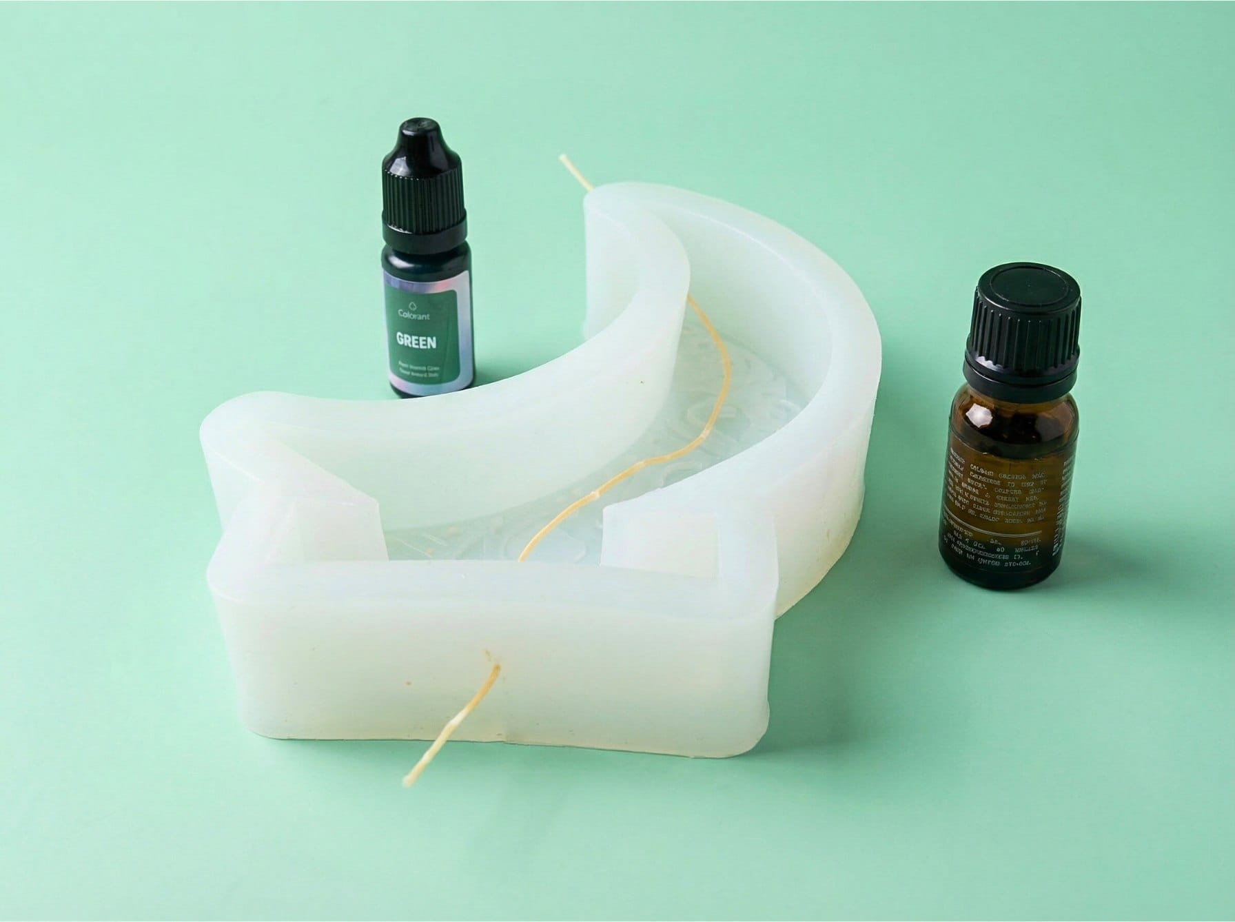 Inserting a cotton wick into a crescent moon silicone mold for a DIY Ramadan beeswax candle, with lavender essential oil and green candle colorant beside it.