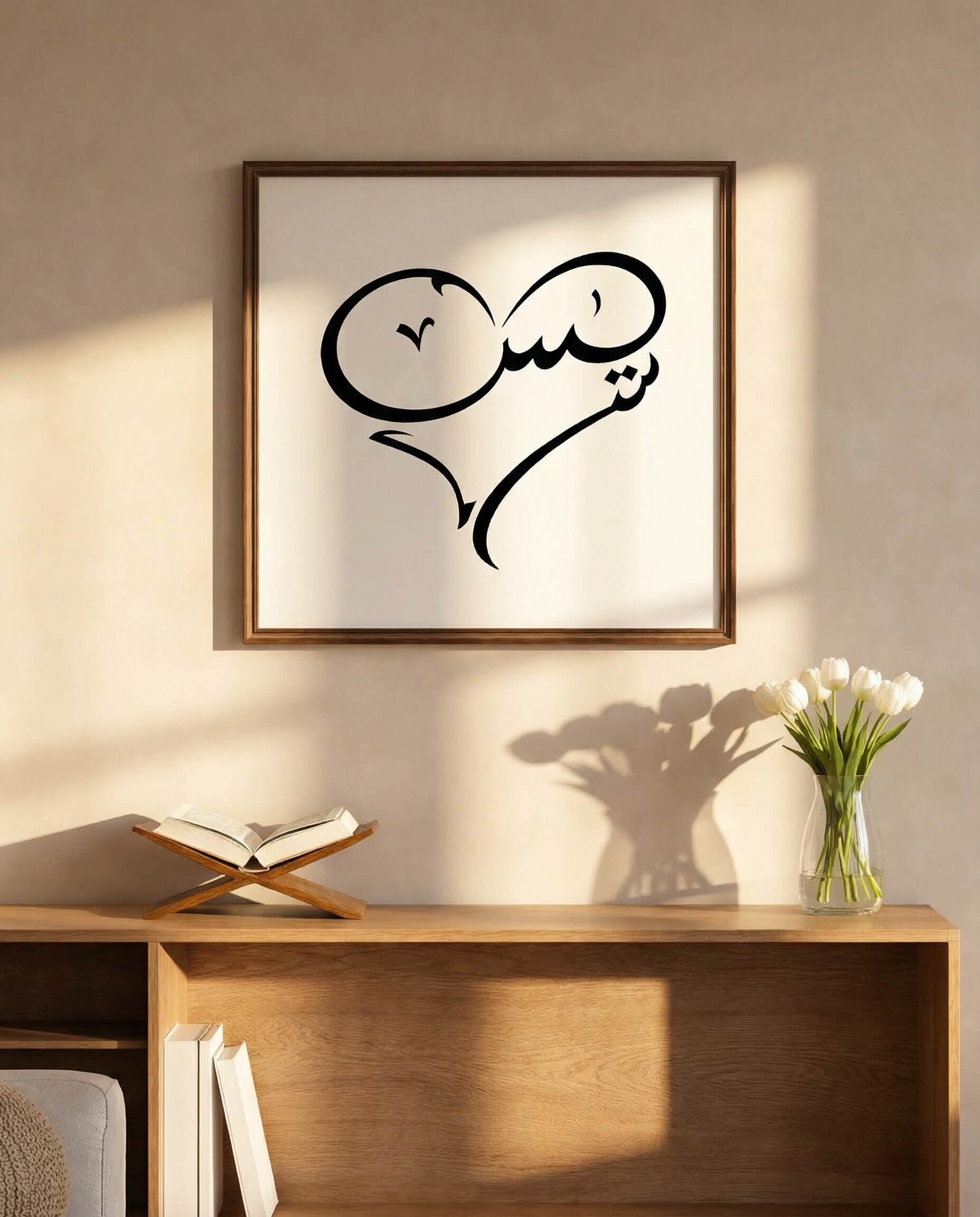 Framed minimalist calligraphy artwork of the word “Yasin” formed into a heart shape, hanging above a wooden console table with an open Quran on a wooden stand.