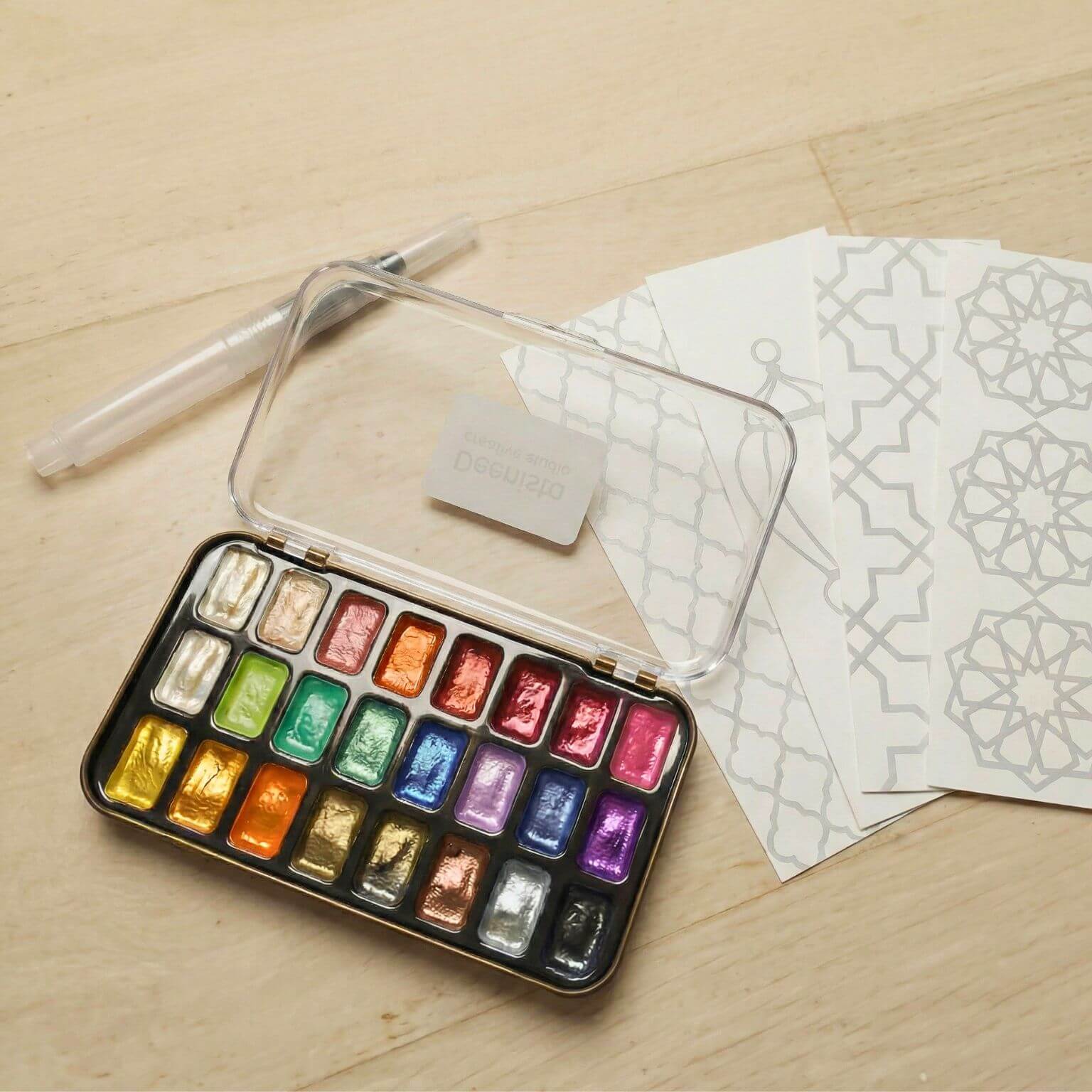 Open Deenista shimmer watercolor kit with a pearlescent paint palette, water brush pen, and four Islamic-pattern bookmark cards ready to paint.