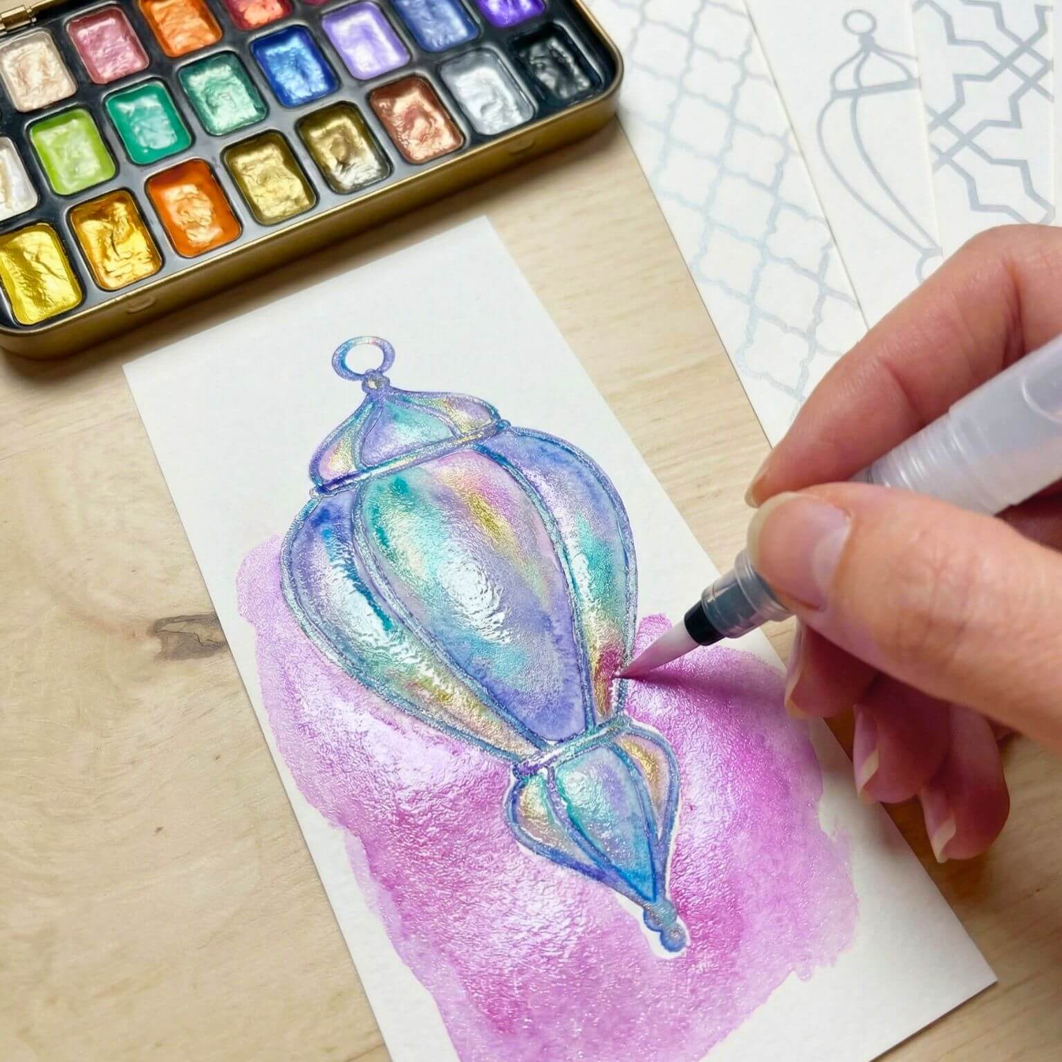 Close-up of a hand-painted Islamic lantern bookmark in shimmering pastel watercolor with pearlescent blue, lilac, pink, and gold tones.
